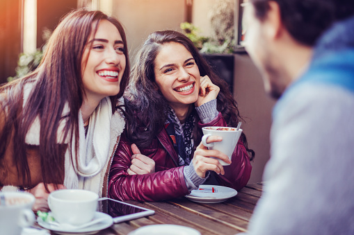 Two young women smiling and talking with a man in a coffee shop.